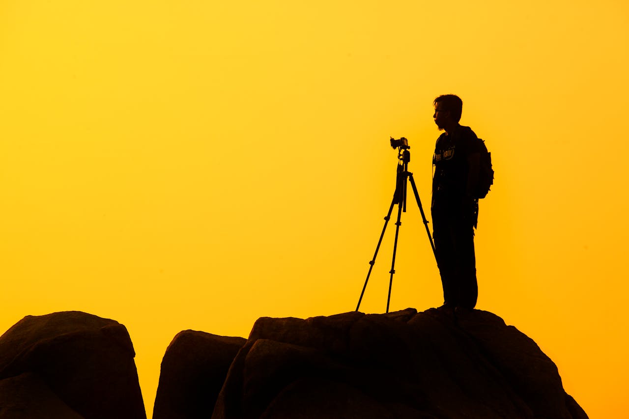 gallery-03 Silhouette of a lone photographer with tripod on a rocky cliff against a golden sunset sky.